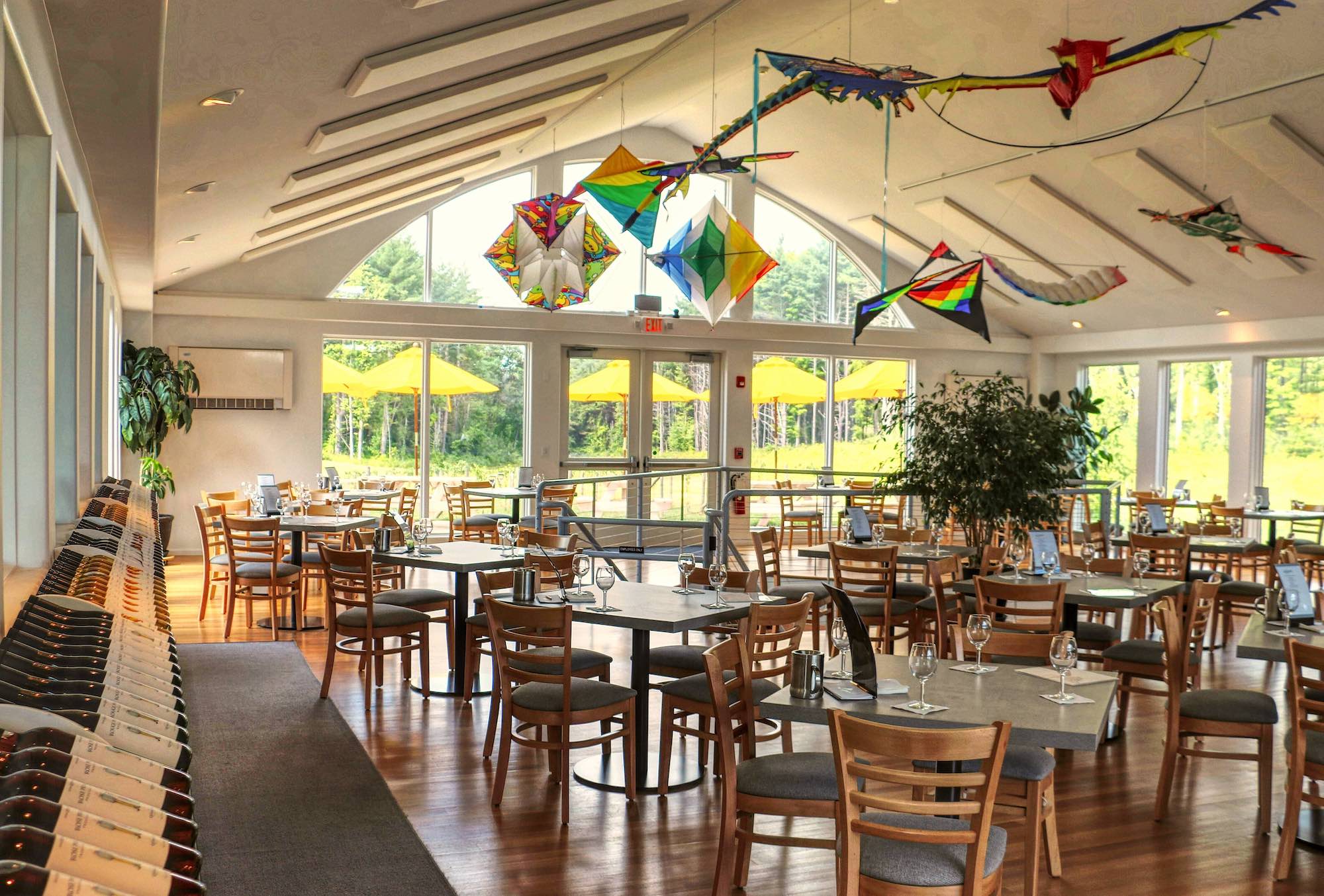Photo of bright, open and airy tasting room interior with tables, chairs and wines arranged in a row on the side. Colorful kits hang from the ceiling and the patio with yellow umbrellas can be seen in window. 
